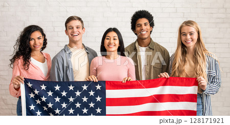 Immigration, relocation and study abroad. Cheerful young international students hold USA flag, looking at camera and ready to learn, on white brick wall background, copy space, studio shot 128171921