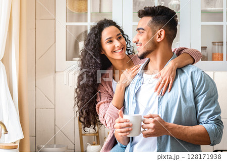 Happy Young Arab Spouses Hugging And Drinking Coffee In Cozy Kitchen Interior. Smiling Millennial Middle Eastern Couple Bonding At Home, Enjoying Spending Morning Time Together, Copy Space 128171938