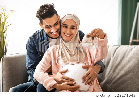 Portrait of happy pregnant muslim couple looking at tiny baby shoes while hugging on couch at home, young loving middle-eastern spouses embracing and enjoying future parenthood, free space 128171945