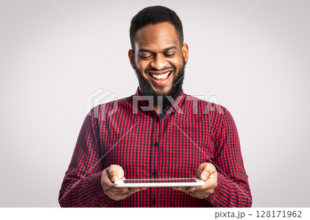 Cheerful African American Man Holding Digital Tablet Standing On White Background. Bearded Black Guy Playing Computer Game Online In Studio. Website, Internet Service And New Application Advert Cheerful African American Man Holding Digital Tablet Standing On White Background. Bearded Black Guy Playing Computer Game Online In Studio. Website, Internet Service And New Application Advert 128171962
