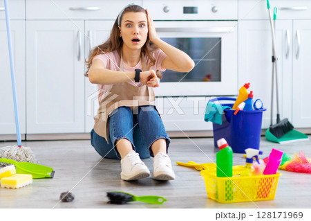 Terrified young housewife looking at her watch, being late with domestic chores on floor at kitchen. Emotional young maid missing housework deadline. Professional cleaning service concept 128171969