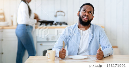 Impatient hungry black husband waiting for dinner with empty plate, bored starving african american man sitting at table and rolling eyes while his wife cooking food on background, free space 128171991