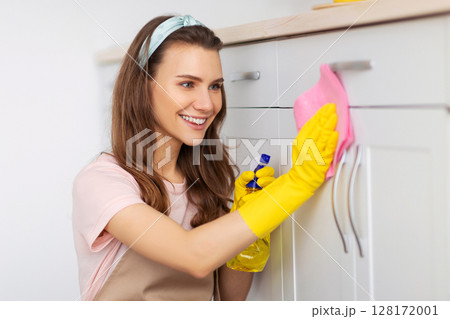 Joyful young woman cleaning kitchen cabinet with detergent and rag, keeping her home tidy. Pretty millennial housemaid doing her professional chores, performing house cleanup 128172001