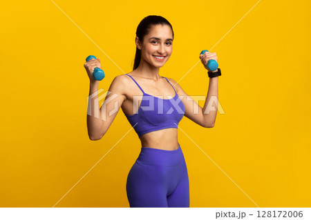 Biceps Exercises, Arms Workout. Portrait Of Happy Young Sportswoman Doing Training With Two Blue Dumbbells Standing Isolated On Orange Studio Background. Smiling Strong Fit Lady In Sportswear 128172006