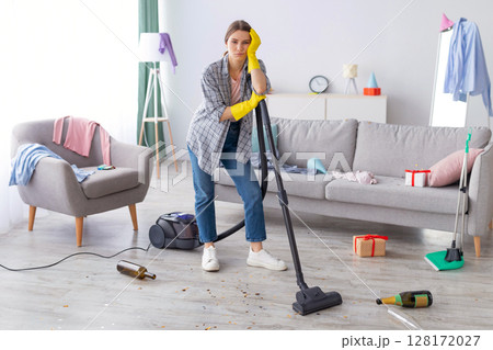 Unhappy young woman standing with vacuum cleaner in messy flat after party, blank space. Upset millennial housewife at dirty room after celebration, garbage scattered everywhere 128172027