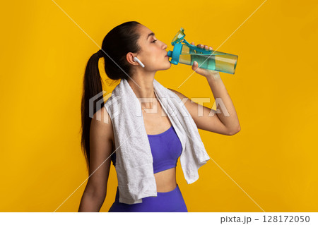 Active Lifestyle. Sporty young lady with white towel on neck drinking mineral water from the bottle, wearing wireless earbuds, listening to workout playlist standing isolated over orange studio wall 128172050