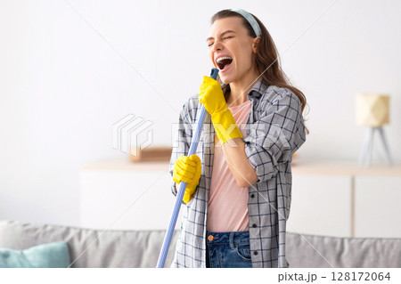 House cleaning is fun. Cheerful young lady using mop as mic, singing song while doing cleanup at living room. Millennial housewife pretending to be rock star while doing domestic duties House cleaning is fun. Cheerful young lady using mop as mic, singing song while doing cleanup at living room. Millennial housewife pretending to be rock star while doing domestic duties 128172064