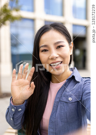 An Asian woman waves hello while taking a selfie outdoors, smiling warmly at the camera. An Asian woman waves hello while taking a selfie outdoors, smiling warmly at the camera. 128172239