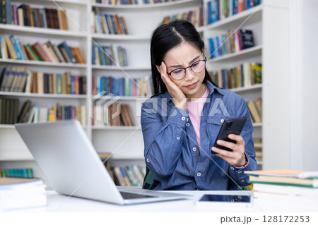 An Asian woman in glasses looks at her phone with a concerned expression in a library, near a laptop. An Asian woman in glasses looks at her phone with a concerned expression in a library, near a laptop. 128172253