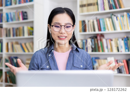 An Asian woman smiles while participating in a video call, using earphones, with a library in the background. 128172262