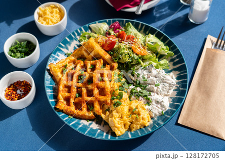 Waffles with scrambled eggs and white cottage cheese served with vegetables and greens on a beautifully decorated plate, a breakfast set, illuminated by the morning sun. 128172705