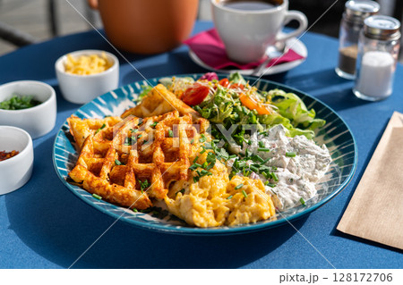 Waffles with scrambled eggs and white cottage cheese served with vegetables and greens on a beautifully decorated plate, a breakfast set, illuminated by the morning sun. Waffles with scrambled eggs and white cottage cheese served with vegetables and greens on a beautifully decorated plate, a breakfast set, illuminated by the morning sun. 128172706