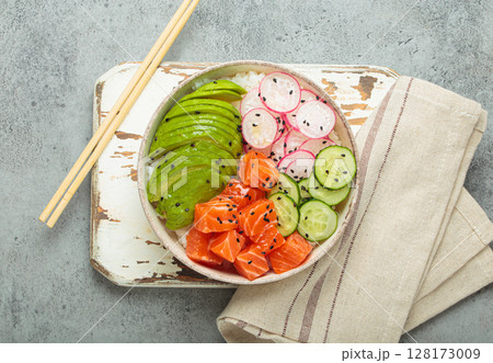 Salmon poke bowl with avocado, radish, cucumber and rice on white rustic wooden board on grey background top view Salmon poke bowl with avocado, radish, cucumber and rice on white rustic wooden board on grey background top view 128173009