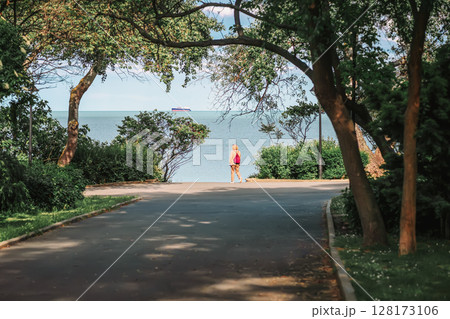 Woman with light backpack walking along green alley in coastal park. Concept of relaxation, sea, travel, active lifestyle, freedom of movement Woman with light backpack walking along green alley in coastal park. Concept of relaxation, sea, travel, active lifestyle, freedom of movement 128173106