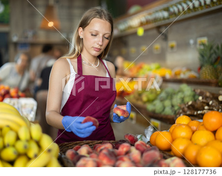 Girl shop seller add flat peach goods on display case in vegetable store Girl shop seller add flat peach goods on display case in vegetable store 128177736