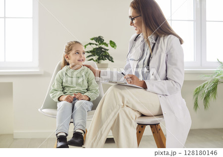 Little girl sitting with smiling doctor during hospital checkup while pediatrician cheering her up. 128180146