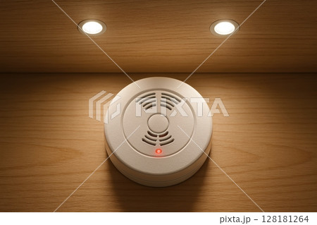 A modern white circular smoke detector with a red indicator light on a wooden ceiling. A modern white circular smoke detector with a red indicator light on a wooden ceiling. 128181264