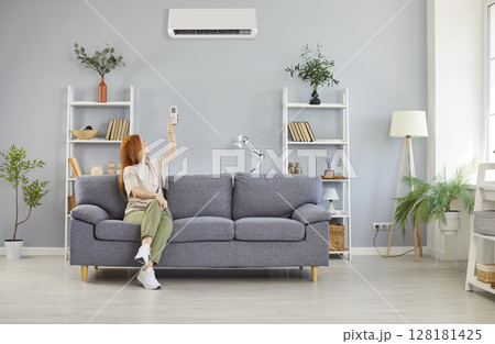 Young woman turning on air conditioner sitting on sofa in the living room at home. Young woman turning on air conditioner sitting on sofa in the living room at home. 128181425