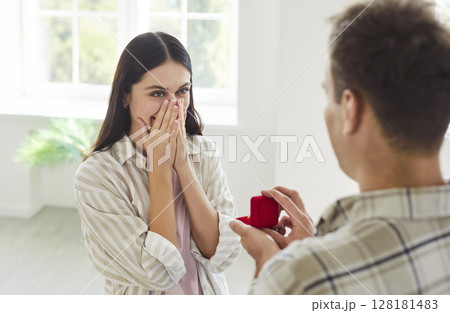 Young man with a red gift box proposing to his happy girlfriend standing in living room at home. Young man with a red gift box proposing to his happy girlfriend standing in living room at home. 128181483