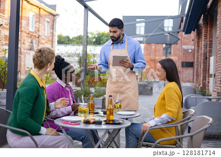 A waiter takes an order on a tablet from customers at an outdoor restaurant. 128181714