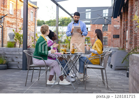 A waiter takes an order from a group of friends sitting at an outdoor cafe table, enjoying drinks and conversation. 128181715
