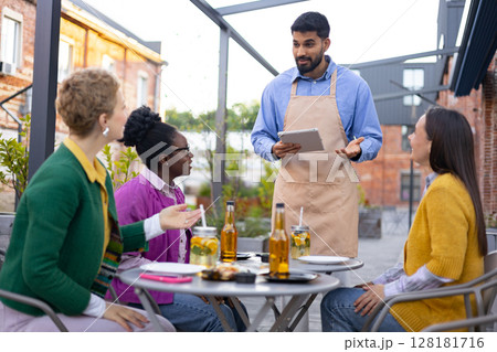 A waiter taking orders from a group of friends at an outdoor cafe, using a tablet device. 128181716