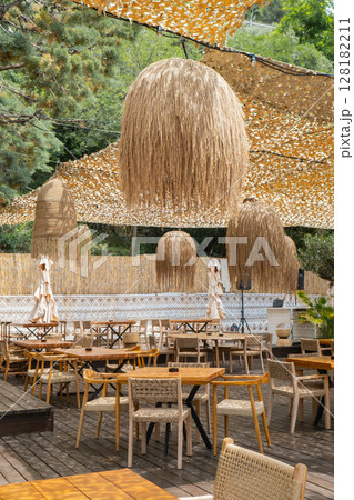 Cozy interior of outdoor cafe on beach seacoast. Wooden tables with thatched bamboo chairs and lamps. Summer terrace with rustic wicker chairs nobody. Natural color rattan furniture restaurant Cozy interior of outdoor cafe on beach seacoast. Wooden tables with thatched bamboo chairs and lamps. Summer terrace with rustic wicker chairs nobody. Natural color rattan furniture restaurant 128182211