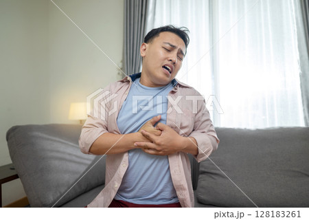 Indonesian southeast asian man lying down on the couch and suffering from chest pain and touching his heart area. Pressure in the chest. Healthcare concept Indonesian southeast asian man lying down on the couch and suffering from chest pain and touching his heart area. Pressure in the chest. Healthcare concept 128183261