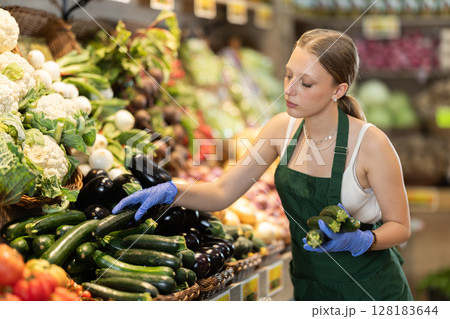 Female salesperson carefully places ripe cucumbers on the counter of grocery supermarket 128183644