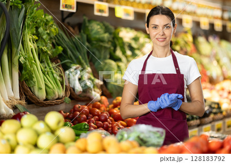 Woman seller in apron and gloves stands in sales area of vegetable store Woman seller in apron and gloves stands in sales area of vegetable store 128183792