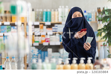 Woman in Muslim attire using phone to check ingredients of body lotion 128183864
