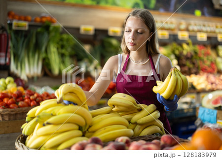 Positive female seller offers to buy ripe tasty bananas in grocery department of supermarket Positive female seller offers to buy ripe tasty bananas in grocery department of supermarket 128183979