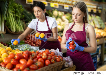 Two girl sellers shop seller add tomato goods on display case in vegetable store 128184029
