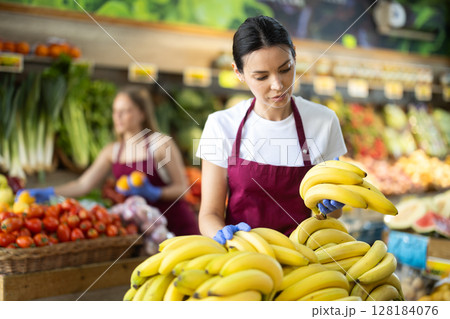 Girl shop seller add banana goods on display case in vegetable store Girl shop seller add banana goods on display case in vegetable store 128184076