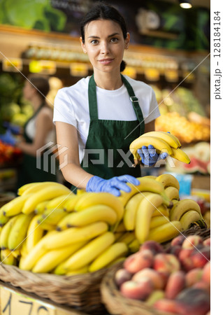 Positive female seller offers to buy ripe tasty bananas in grocery department of supermarket 128184184