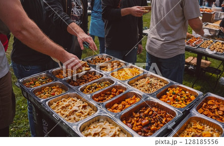 Group of people are gathered around a table with a variety of food, including a tray of macaroni and cheese Group of people are gathered around a table with a variety of food, including a tray of macaroni and cheese 128185556