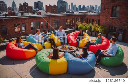 Group of people are sitting on colorful bean bags on a rooftop. Scene is relaxed and social, as the group of people are gathered together to enjoy each other's company. The colorful bean bags add a 128186740