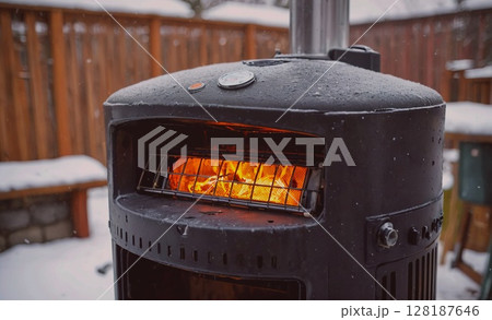 Black outdoor heater with a fire burning inside. The fire is warm and inviting, and the heater is surrounded by snow Black outdoor heater with a fire burning inside. The fire is warm and inviting, and the heater is surrounded by snow 128187646