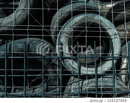 A Stack of Used Tires Behind a Metal Fence in Direct Sunlight 128187988