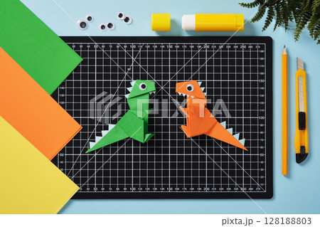 Green and orange origami dinosaurs on a black cutting mat. Surrounded by colorful sheets of paper, googly eyes, glue stick, pencil, and craft knife. DIY project concept for kids. 128188803