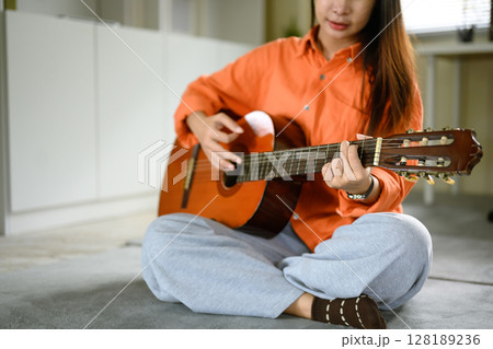 Asian female playing guitar indoors with natural light streaming through the blinds 128189236