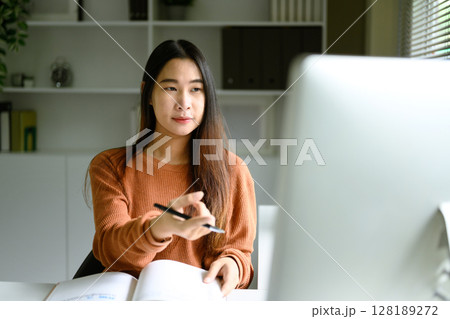 Young woman in a warm toned sweater working at a desktop computer in a home office 128189272