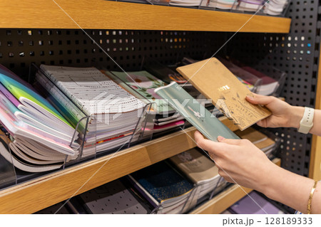 A person selects a notebook with NOTE printed on its cover from a diverse stationery shelf in a retail store, symbolizing shopping for school or office supplies 128189333