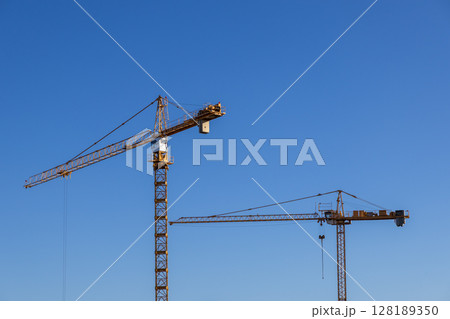 Two large yellow tower cranes stand tall against a clear blue sky, symbolizing active construction, urban development, and industrial infrastructure growth Two large yellow tower cranes stand tall against a clear blue sky, symbolizing active construction, urban development, and industrial infrastructure growth 128189350