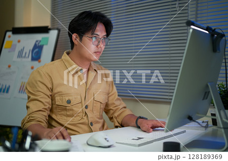 Focused man in glasses working late on desktop computer in a dimly lit office Focused man in glasses working late on desktop computer in a dimly lit office 128189369