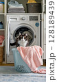 A playful cat is peeking out from inside an open washing machine, next to a laundry basket with clothes. 128189439