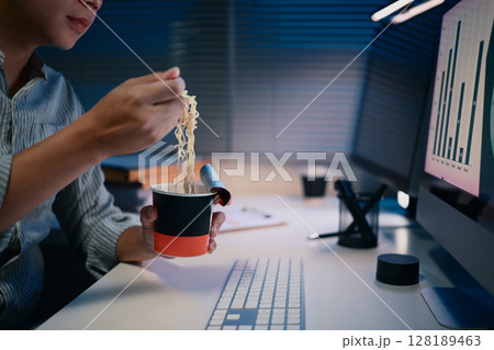 Close up of man eating instant noodles at desk while working late at night under office lighting Close up of man eating instant noodles at desk while working late at night under office lighting 128189463