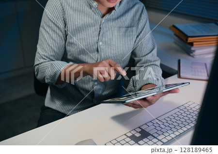 Man using digital tablet while working late at night at a desk with documents Man using digital tablet while working late at night at a desk with documents 128189465