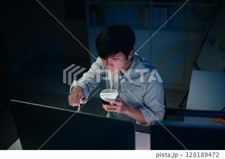 Man eating instant noodles while working on a computer late at night in a dim office Man eating instant noodles while working on a computer late at night in a dim office 128189472