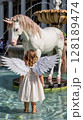 A charming little girl with angel wings stands before a magical unicorn statue in a fountain. 128189474